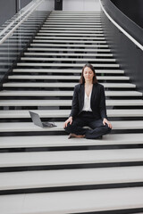 business woman in a formal suit with a laptop in the office of the business center sits on the steps in the lotus position and meditates, relax. Work in a corporation as a marketing financier or