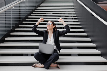 business woman in a strict suit with a laptop in the office of the business center is sitting on the steps rejoicing at a successful contract victory. Work in a corporation as a marketing financier or