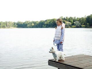 A seven-year-old girl with a small white dog stands on a bridge near the lake and looks at the water. Friendship with pets. Happy childhood.