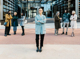 Successful businesswoman standing with crossed arms outside the office building in financial district with teamwork on the background. Female entrepreneur, business and success concept.