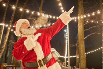 Waist up portrait of Santa Claus dancing outdoors at night and blowing smoke, copy space
