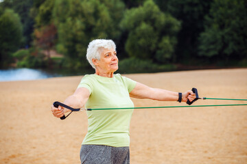 Old woman wear sportswear doing exercises outdoor on the beach in the morning using resistance rubber bands. Healthy lifestyle, active retired life and sporty time.