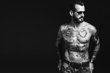 Fototapeta na wymiar Portrait of handsome confident stylish hipster lambersexual model. Sexy modern man. Naked torso with tattoos.Fashion male posing in studio on dark background. Black and white