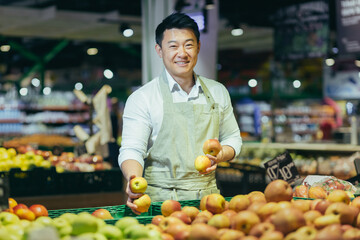 Waist up portrait of a handsome Asian handsome man working in a supermarket and smiling at the camera. Standing in a work apron near separated vegetables and fruits. He holds apples in his hands.