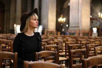 Woman in black dress and hat sits alone in Catholic church. Pray time