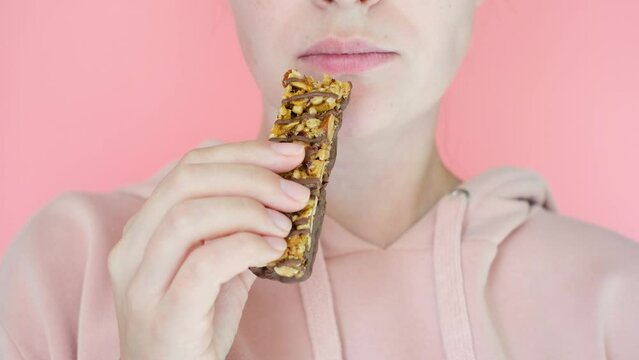 Young woman eating protein bar on pink background close-up.