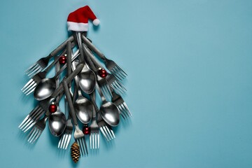 Christmas tree made of spoons and forks with red berries in Santa Claus hat on a light blue background. Top view with copy space. Christmas creative composition. Card or menu template