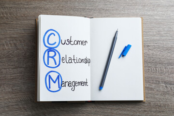 Notebook with abbreviation CRM (Customer Relationship Management) and pen on wooden background, top...