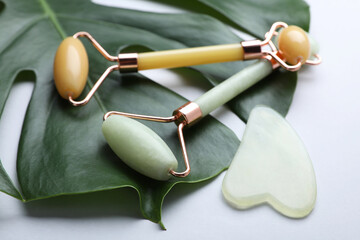 Gua sha stone, different face rollers and monstera leaf on light background