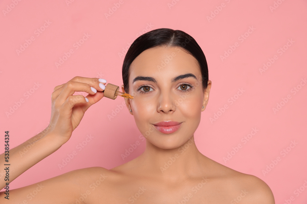 Wall mural Young woman applying essential oil onto face against pink background