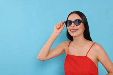 Obraz premium Attractive happy woman touching fashionable sunglasses against light blue background. Space for text