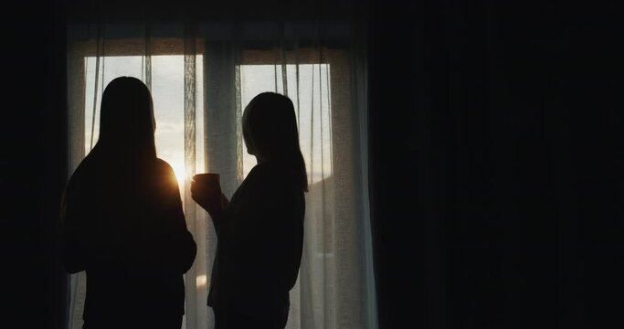 A woman with a teenage daughter stand at the window at sunset. Holding cups of tea. Rear view
