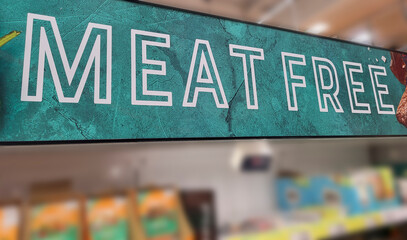 Close up color image depicting a Meat Free sign indicating the vegetarian food section inside a supermarket. Focus is on the sign, while the aisle of the supermarket is entirely defocused.