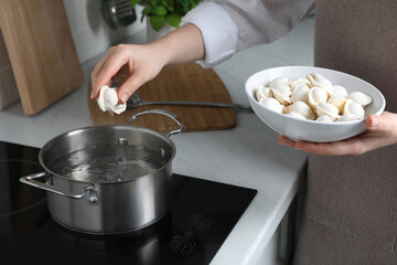 Woman putting frozen dumplings into saucepan with boiling water on cooktop in kitchen, closeup