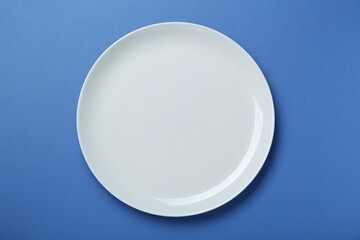 Empty white ceramic plate on blue background, top view