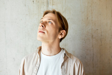 Studio portrait of handsome caucasian hipster guy with blond hair and blue eyes standing on gray textured wall looking up at something, dressed in casual beige shirt and white t-shirt