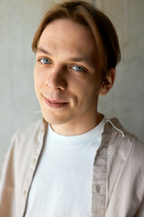 Vertical studio portrait of cute charming handsome caucasian man with blond hair and stylish haircut looking at camera smiling on gray textured wall, wearing casual beige shirt upon white t-shirt