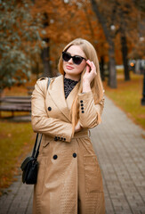  portrait of blonde woman in sunglasses  and in beige coat walking  in autumn park