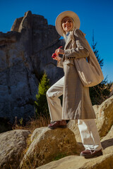 Happy smiling fashionable woman wearing stylish autumn outfit with white hat, wide jeans, midi coat, beige leather hobo bag, posing in mountain nature. Full-length outdoor portrait