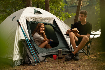 Shot of man hiker with his girlfriend reading book and camping in summer wood.