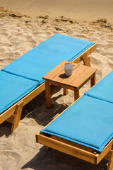 Closeup view of two blue sunbeds and a small table in front of the beautiful golden sandy beach of Mylopotas in Ios Greece