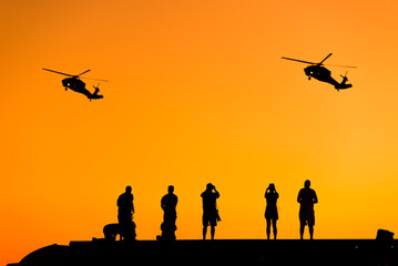 Naklejka na ściany i meble Silhouette of some photographer people taking photo of helicopters on sunset.
