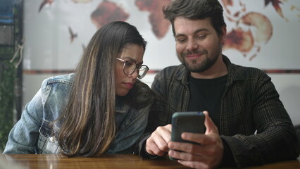 Couple looking at smartphone screen seated at coffee shop. People laughing and smiling over video content online. Young man showing phone screen to girl