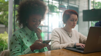 People using cellphone and laptop at coffee shop. Two young women staring at screens. Girl looking at computer browsing internet inside cafe restaurant