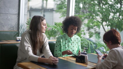 Fototapeta premium Candid group of friend seated at coffee shop. Diverse women at cafe restaurant. Three girlfriends at restaurant looking at gadgets