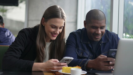 Fototapeta na wymiar An interracial couple looking at cellphones seated at coffee shop. Diverse people smiling using smartphone device. Girlfriend with boyfriend at cafe place