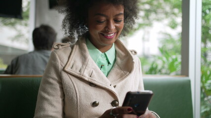 One happy black woman checking phone at coffee shop. Smiling African American girl with Afro hair reading cellphone message