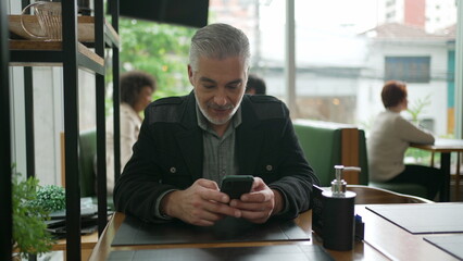 Older man using cellphone inside coffee shop watching content online on phone. A middle aged person...