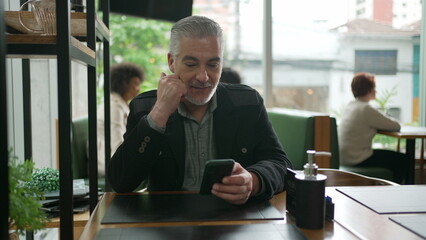 Older man using cellphone inside coffee shop watching content online on phone. A middle aged person looking at smartphone screen