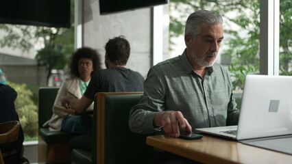 Middle aged man preparing to work adjusting shirt and organizing coat and items in front of laptop seated at coffee shop
