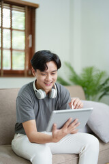 A portrait of a young man showing a smiling face while relaxing, using a tablet computer and a pair of headphones to listen to music