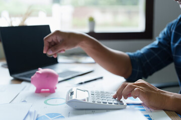 Portrait of an Asian businessman using a calculator to calculate his savings from SME operations, with a pink piggy bank as keep money concept