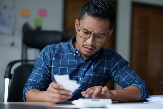 Portrait of a male SME business owner with a stressed and anxious face using a calculator to calculate expenses from the bill on hand and annual income tax to pay the Revenue Department