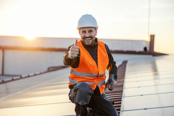 A happy worker with drill in his hands is giving thumbs up for using solar panels while holding a drill and smiling at the camera. - obrazy, fototapety, plakaty