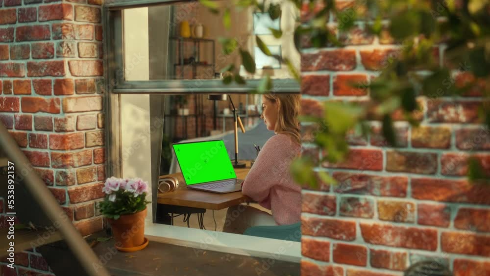 Fototapete Female Working on a Laptop Computer with Green Screen Mock Up Display while Sitting Behind a Table in Stylish Living Room. Business Manager Working Remotely from Home. View from Outside the Window.