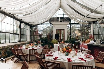 decorated greenhouse hall for a festive event with white capes and country chairs. numbered and decorated tables in a room with many windows and no people.
