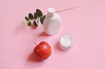 White shampoo bottle, moisturizing cream, fresh apple, eucalyptus branch on a pink table top view photography. Natural organic cosmetics for hair and skin care