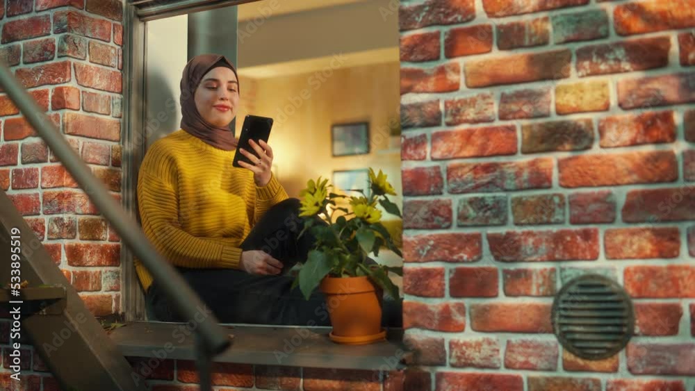 Aufkleber young muslim female using smartphone while sitting on a window sill in a multi-storey brick house. c