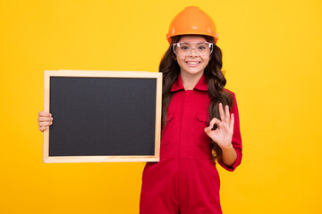 Child girl builder in hard hat helmet. Teenage girl worker hold blackboard isolated on yellow background. Kids renovation concept. Copy space, mock up.