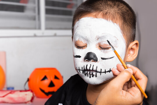 Asian mother and kid son getting makeup halloween face painting look like ghost, Portrait of woman applying color brush to child face for party, Happy halloween day concept