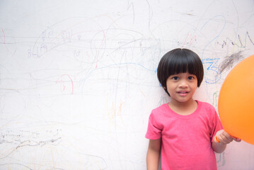 A smiling Asian youngster carrying an orange balloon while donning a pink outfit. On the house's white wall, he painted with several crayons. copy space.