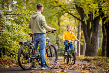 Happy father and daughter ride bicycles in autumn park on sunny day.