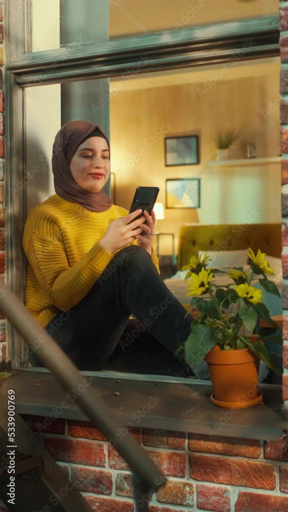 Poster vertical screen: muslim girl wearing a hijab, using a smartphone while sitting on a window sill in b