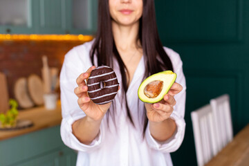 a young woman in a white shirt stands in the kitchen with a donut in one hand and an avocado in the other hand, makes a choice