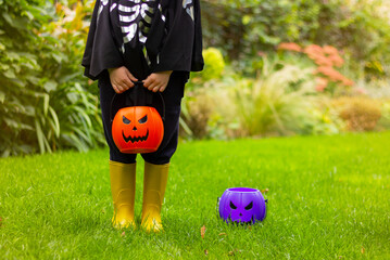 child in black halloween bat costume and yellow boots on lawn holding pumpkin buckets for candy, no face