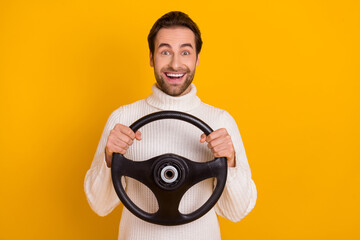 Photo of cute excited guy dressed white sweater holding steering wheel riding car isolated yellow...
